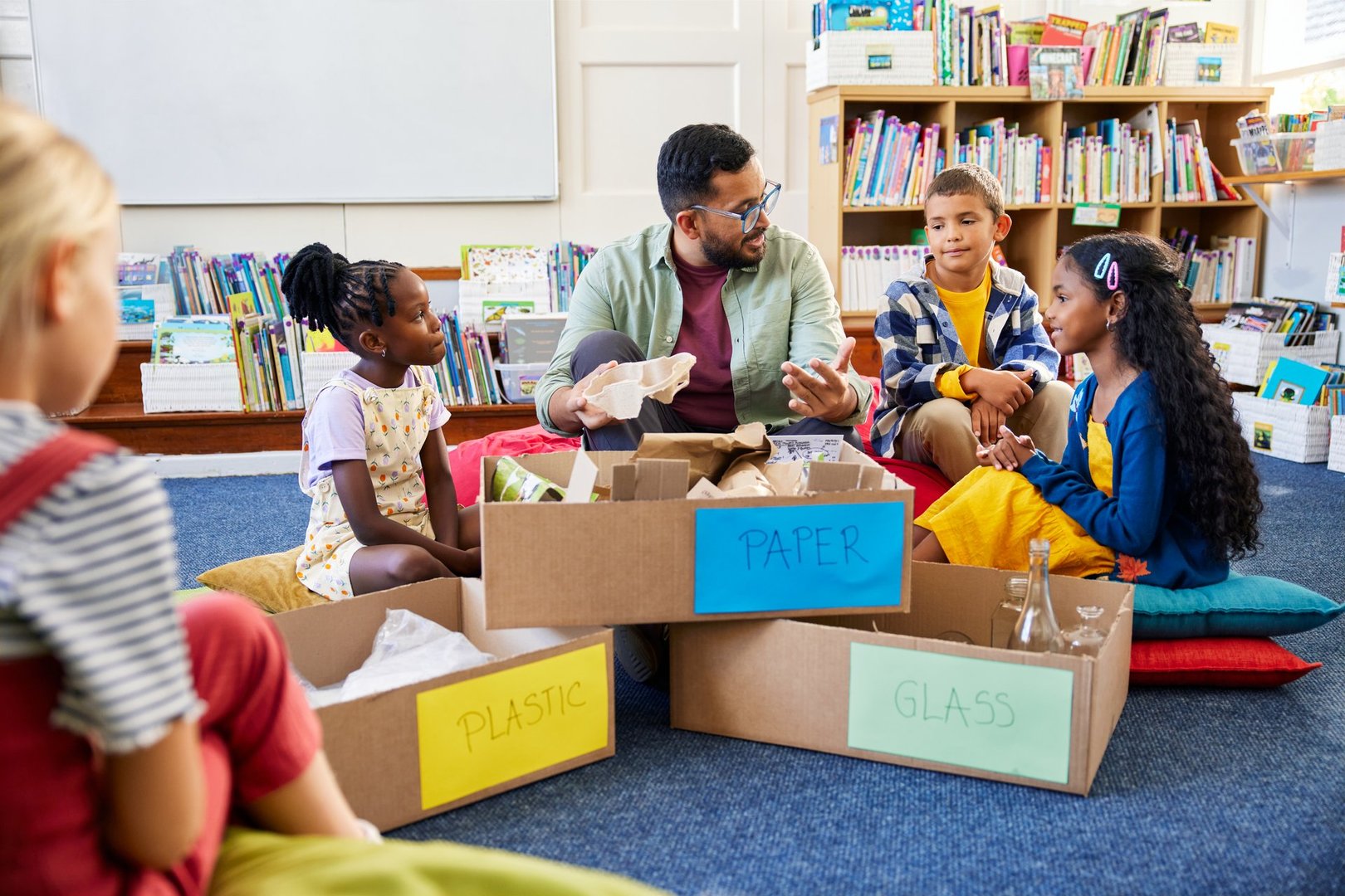 Group of kids with male teacher organizing paper, plastic and glass in cardboard boxes in classroom to throw them away separately. Teacher educates the elementary class in separating waste: paper, plastic and glass for recycling. Multiethnic children participate in the separation of plastic, paper and glass waste to save the planet from pollution.