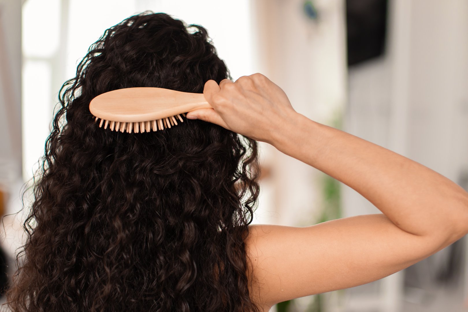 Back view of young brunette woman brushing her long wavy hair with wooden brush at home. Millennial female taking care of her beautiful curly locks indoors. Domestic beauty routines concept