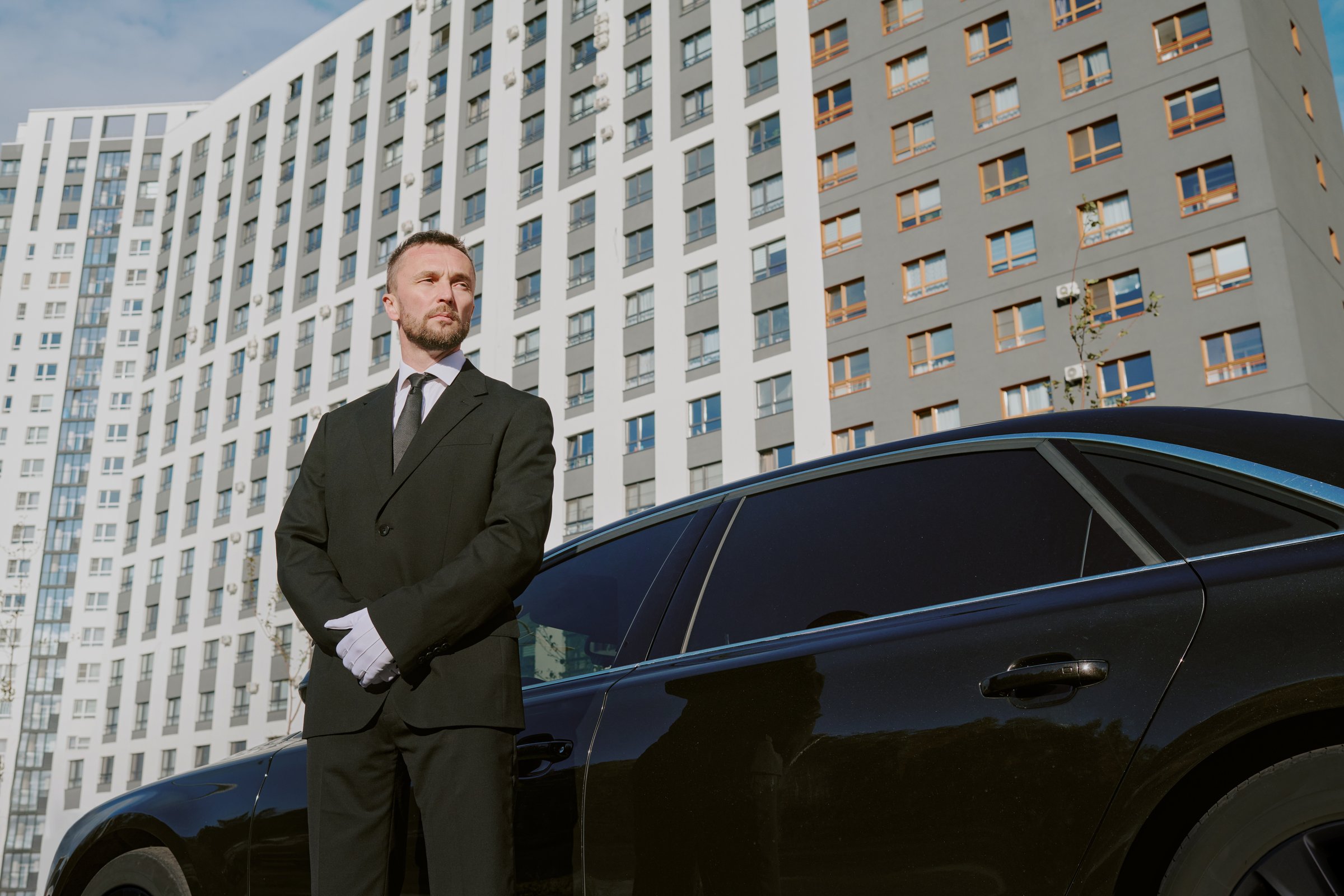 Caucasian middle aged man standing in front of black car outside modern apartment building, wearing suit and gloves, looking away with serious expression, acting as security guard