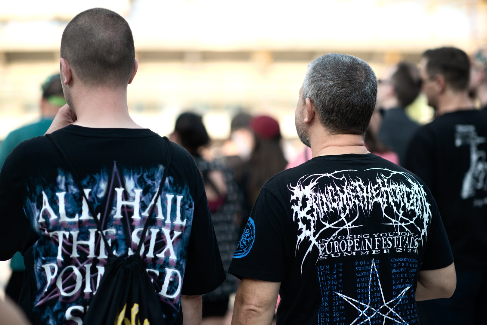 Plovdiv, Bulgaria - July 25, 2025:Two men dressed in black metal band T-shirts and casual clothing stand in a live outdoor music festival crowd during a heavy metal concert, surrounded by other attendees in daylight atmosphere.