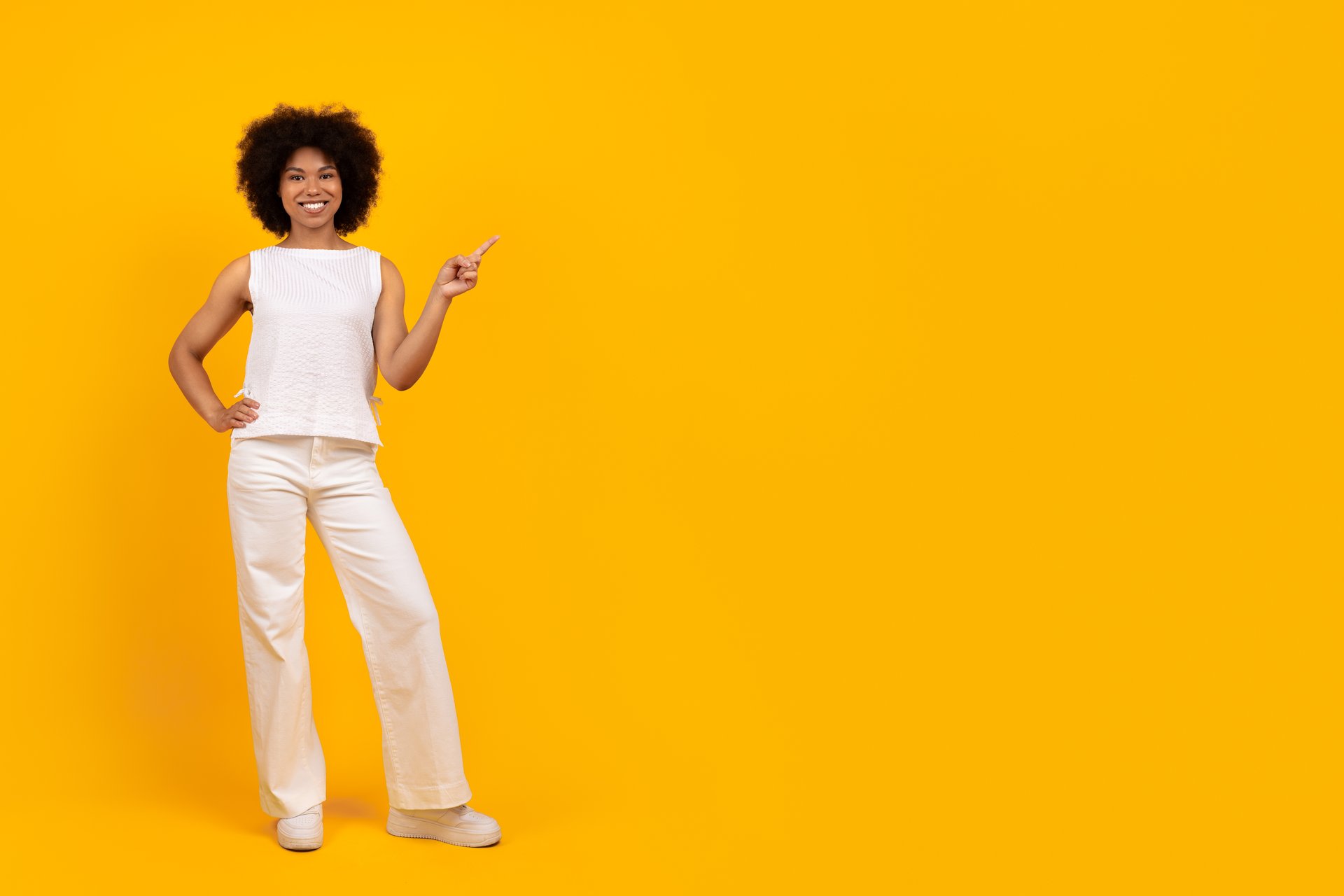 Check This. Positive cheerful beautiful young african american female in white outfit showing copy space for advertisement, happy black female standing over yellow studio background, full length shot
