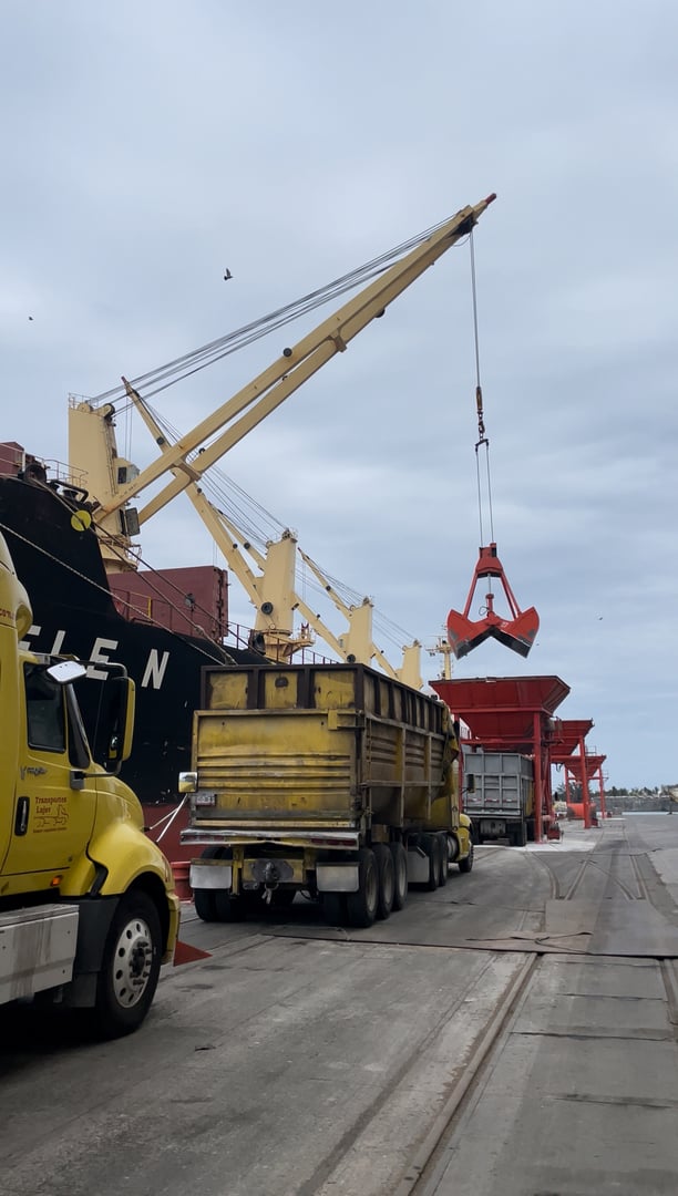 A crane unloads cargo from a ship into a large yellow truck at a dock, with a cloudy sky in the background.
