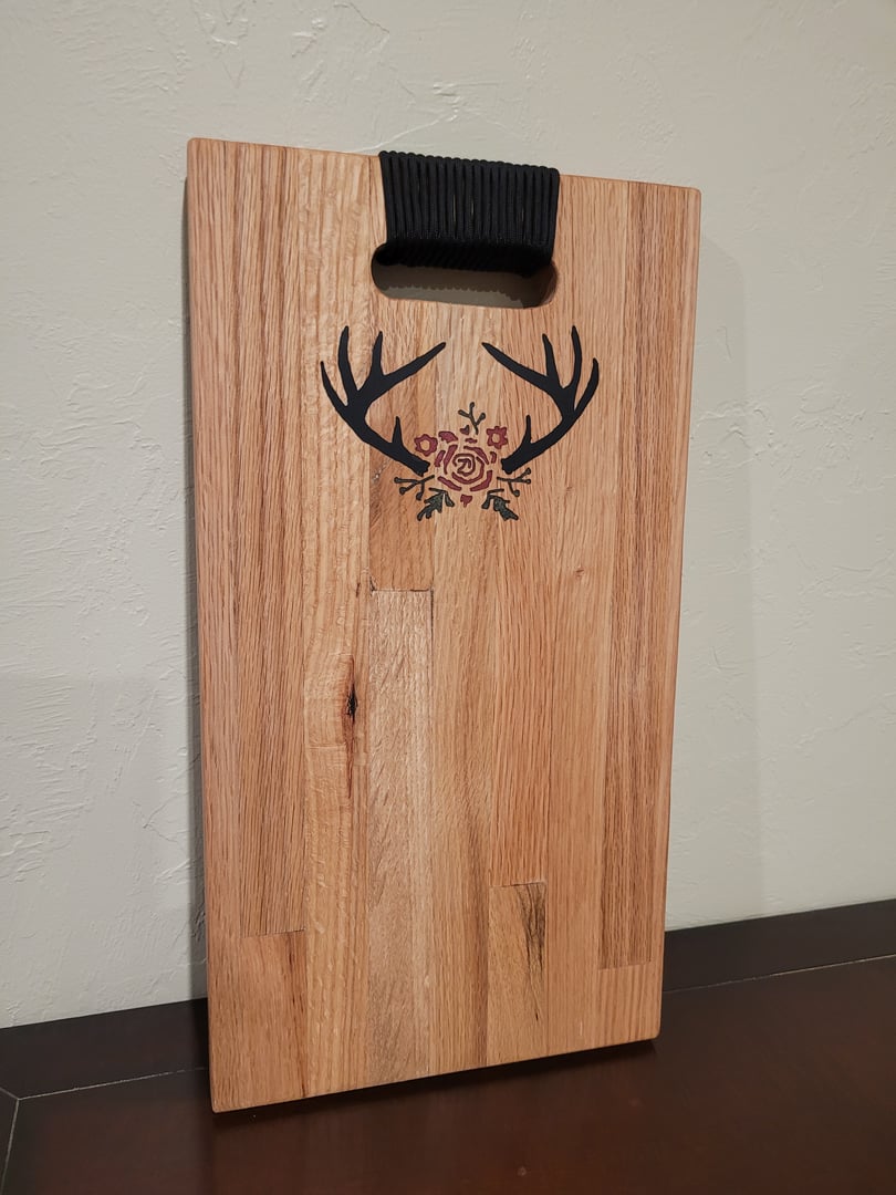 Wooden cutting board with antler and floral design, featuring a black rope handle.