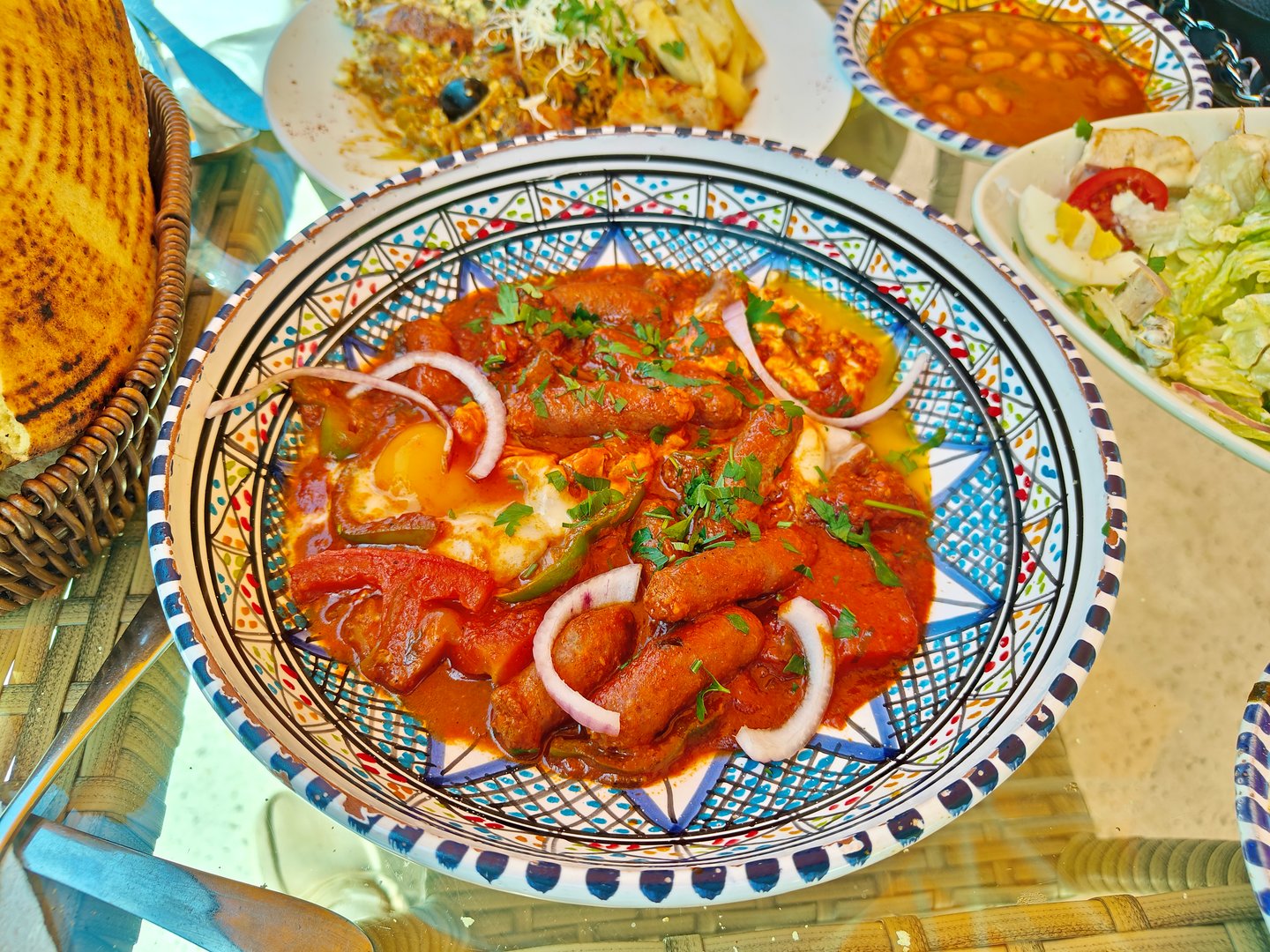 A vibrant and flavorful Tunisian dish called "Ojja" featuring spicy merguez sausages, poached eggs, bell peppers, onions, and tomato sauce, served in a traditional ceramic bowl. Accompanied by various side dishes and fresh flatbread.