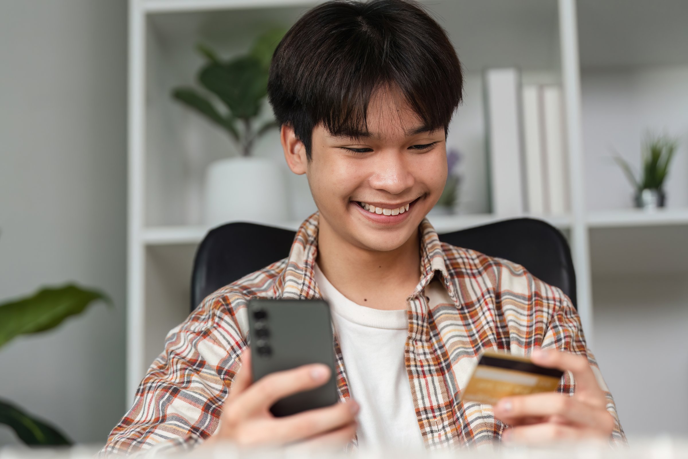 A young adult expresses excitement while managing a financial transaction using a smartphone and credit card.