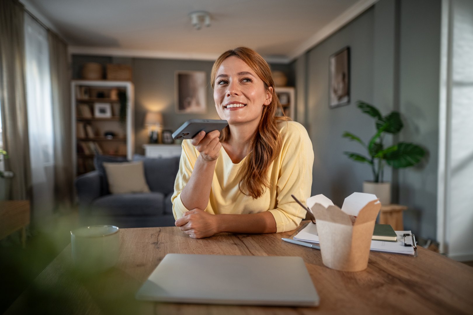 Woman comfortably sitting at a wooden table in her living room, smiling and holding a smartphone near her mouth, recording a voice message or using a voice assistant application