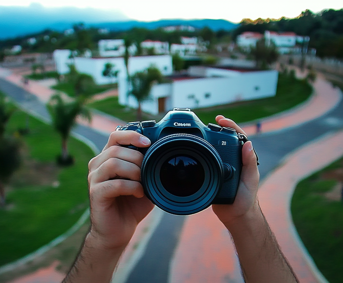 Person holding a professional camera in front of a blurred background of a winding path and white buildings.