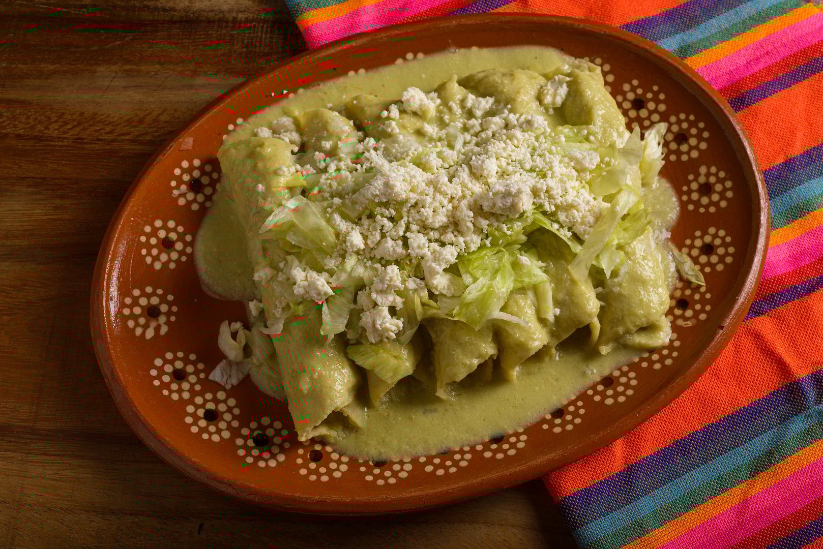 Green enchiladas with lettuce and cheese served in a clay dish. Colorful tablecloth. Close up