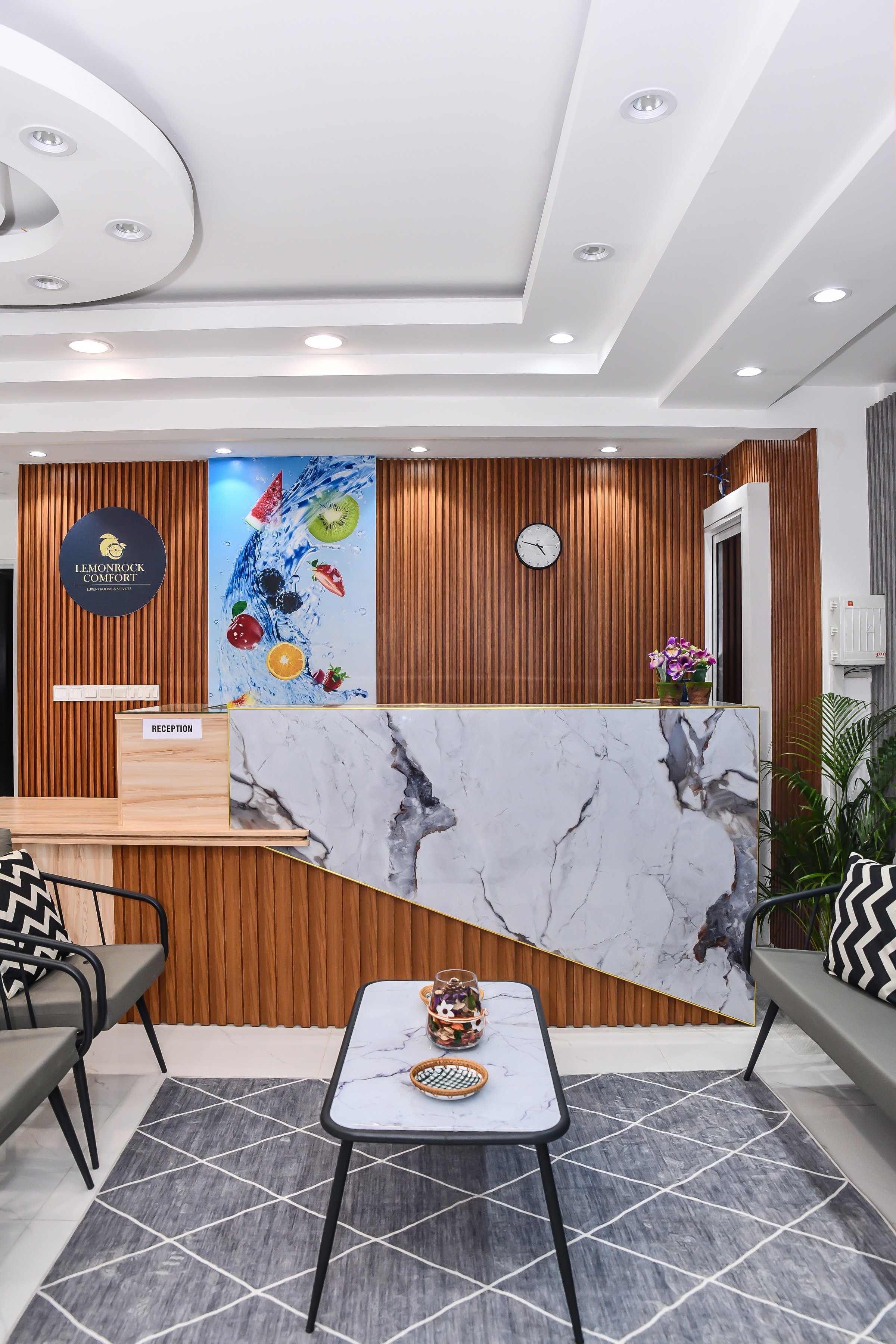 Modern reception area with marble desk, wooden paneling, wall clock, and contemporary decor including plants and artwork.