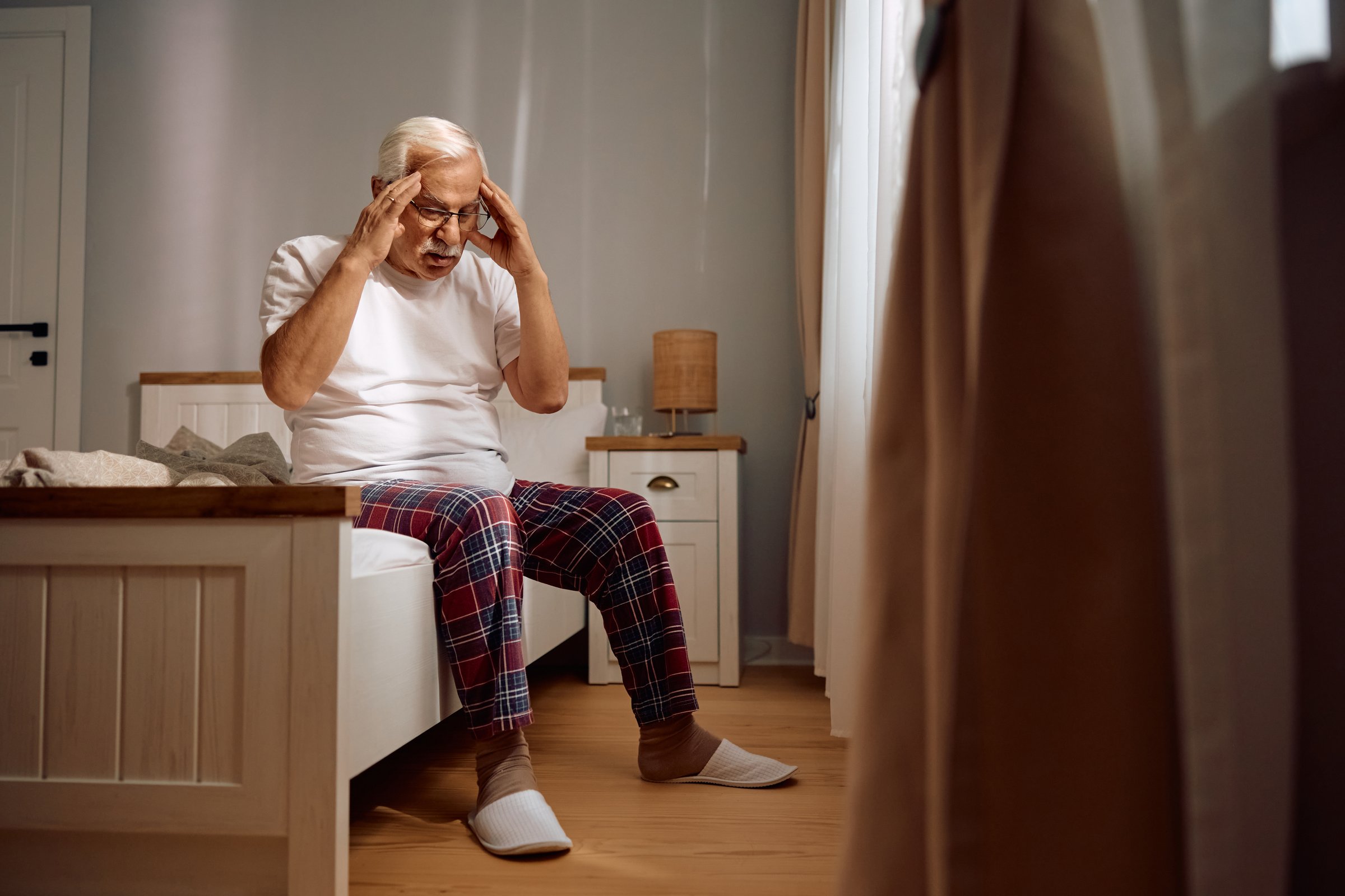 Senior man holding his head in pain while sitting on a bed at home. Copy space.