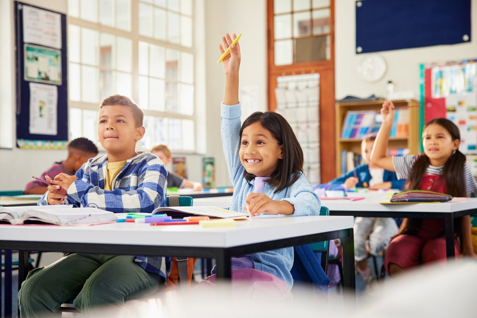 Excited hispanic little girl raising hand in elementary classroom. School children during the lesson trying to answer question in primary school. Cute little latin girl raising hand at school with funny expression.
