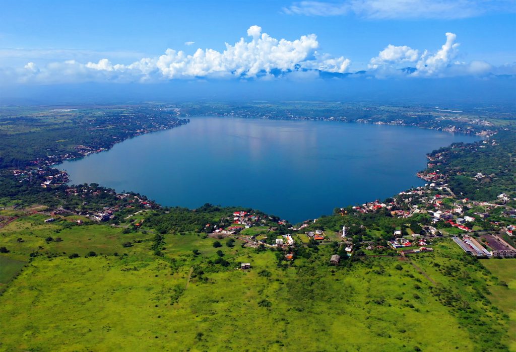 Aerial view of a large lake surrounded by lush green landscape and scattered houses under a partly cloudy blue sky.