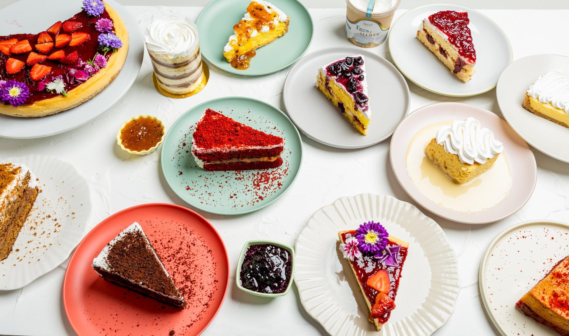 Assorted slices of cake on plates, including strawberry, red velvet, and chocolate, with decorative flowers and toppings.