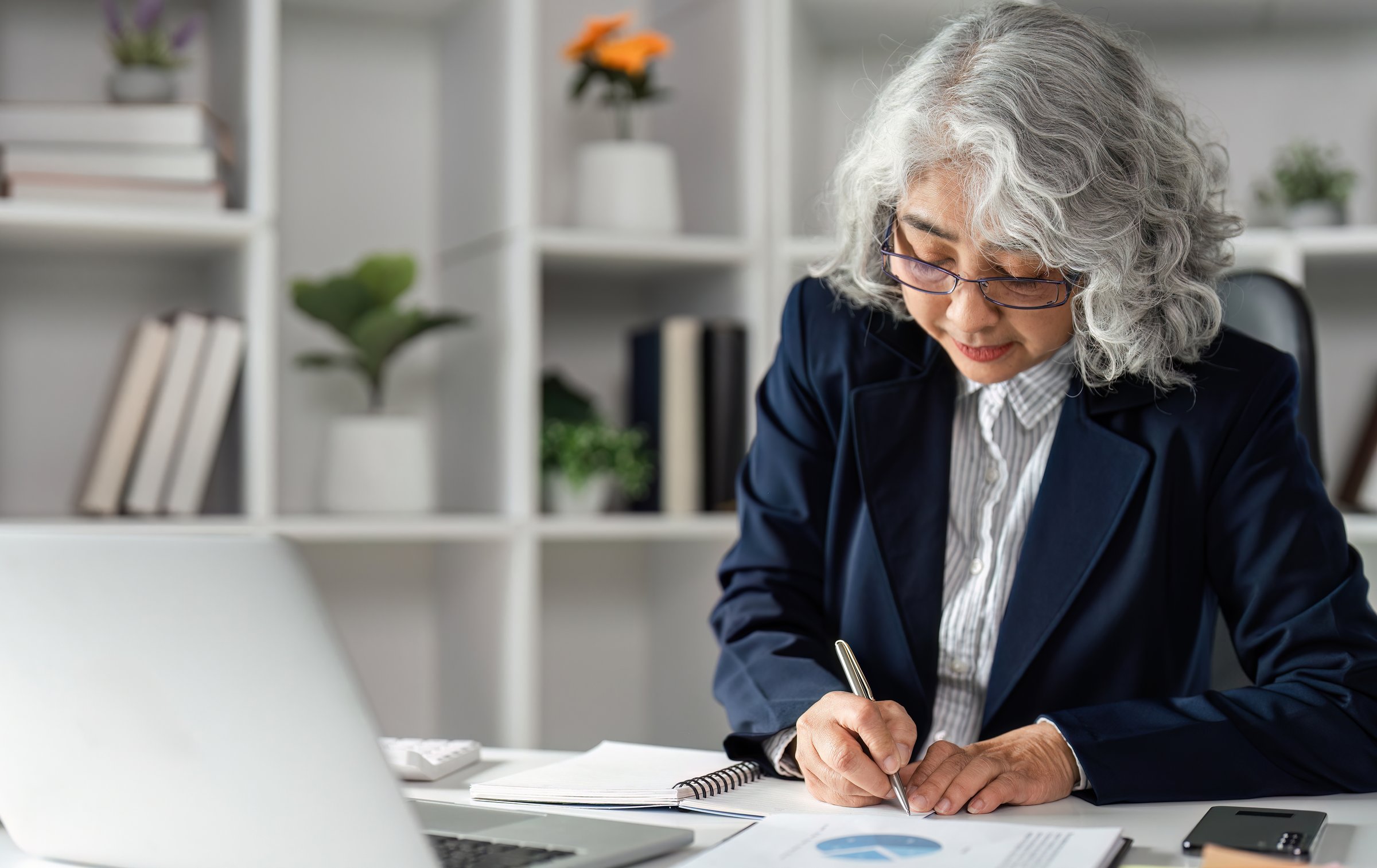 An elderly business woman is focused as she takes notes and analyzes data charts at her contemporary desk, highlighting her dedication and professionalism.