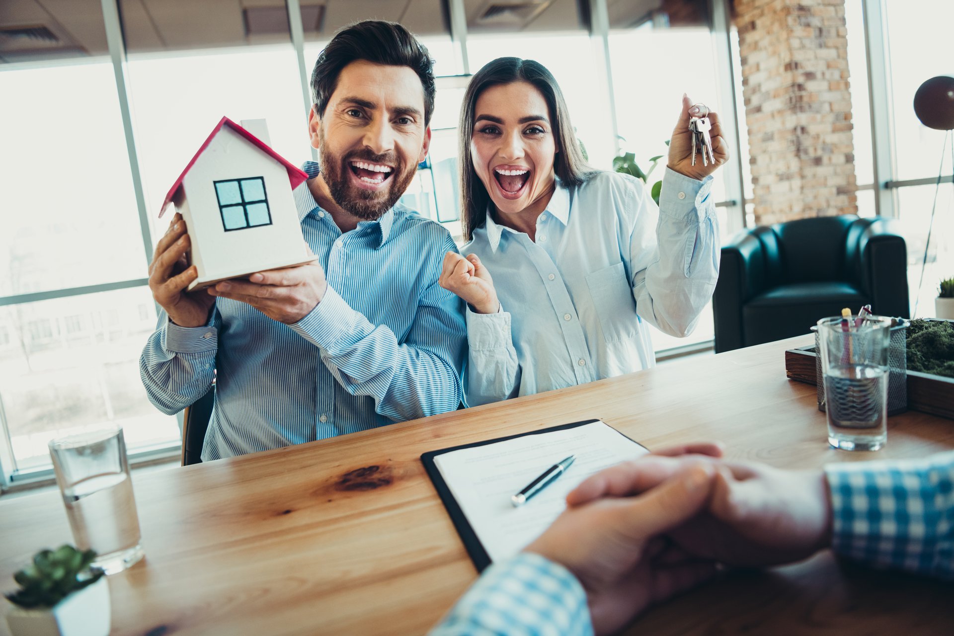 A happy couple at a business office excitedly hold house keys and a model house, symbolizing their new home ownership success