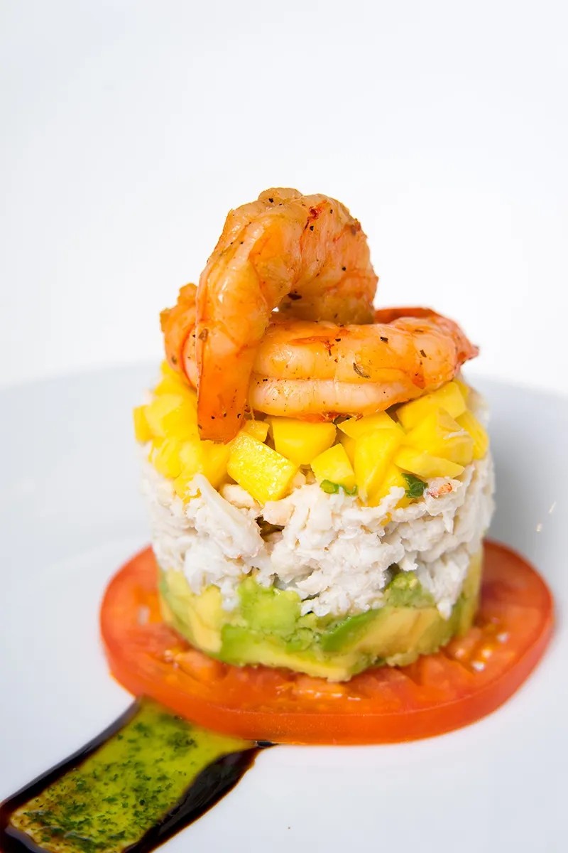 Layered seafood dish with shrimp, mango, crab, avocado on tomato slice, topped with sauce on a white plate.