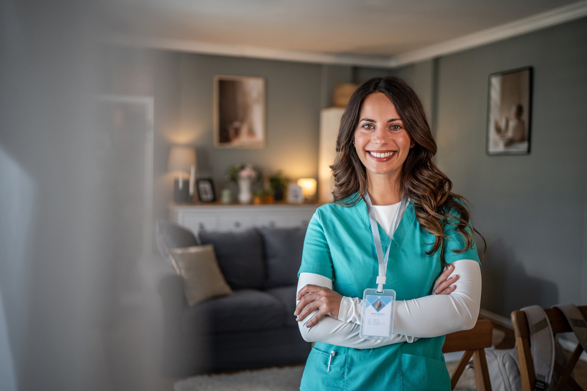 Professional female healthcare worker in scrubs standing with arms crossed, smiling at the camera, providing home care in a comfortable living room setting