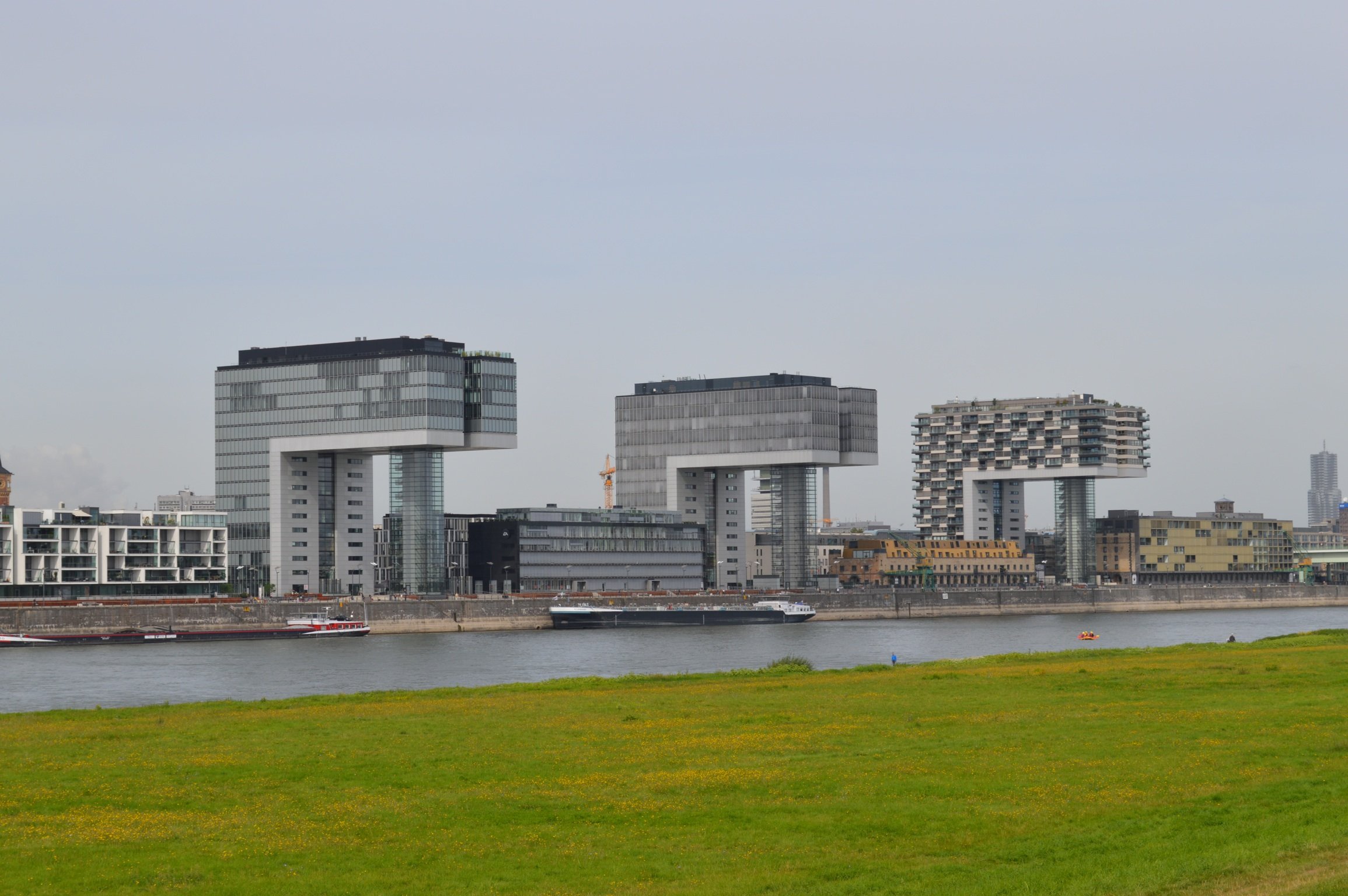 Three modern crane-like buildings by a river, with grassy foreground and city skyline in the background.