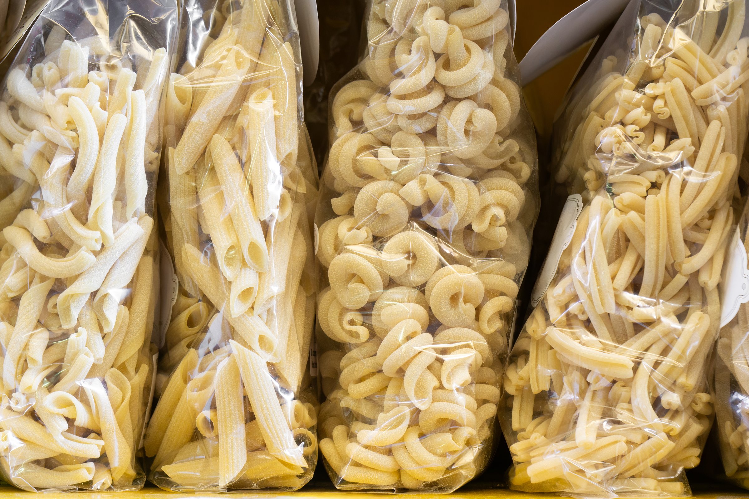 Various types of italian pasta, including penne and casarecce, are displayed in clear plastic bags on a store shelf, ready for purchase
