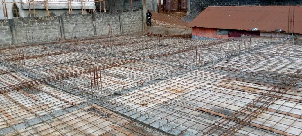 Construction site with rebar grid on concrete slab, adjacent to a corrugated metal-roofed building and gravel pile.