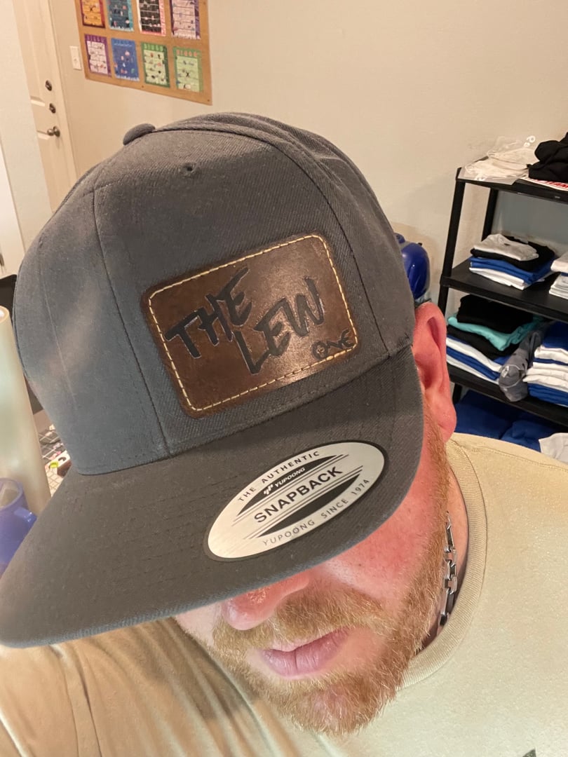 Person wearing a black cap with THE LEW on a brown leather patch. Cap shows SNAPBACK YUPOONG. Background has shelves.