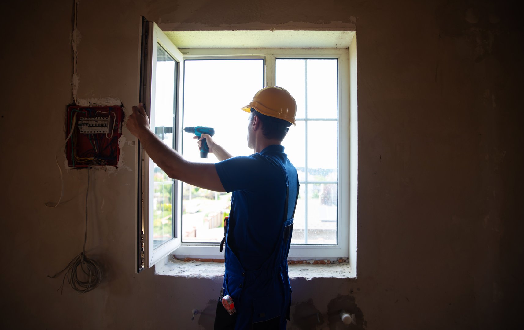 A construction worker in a yellow hard hat installs or adjusts a window frame with a drill in an unfinished room. The open window provides a view of the outdoors.