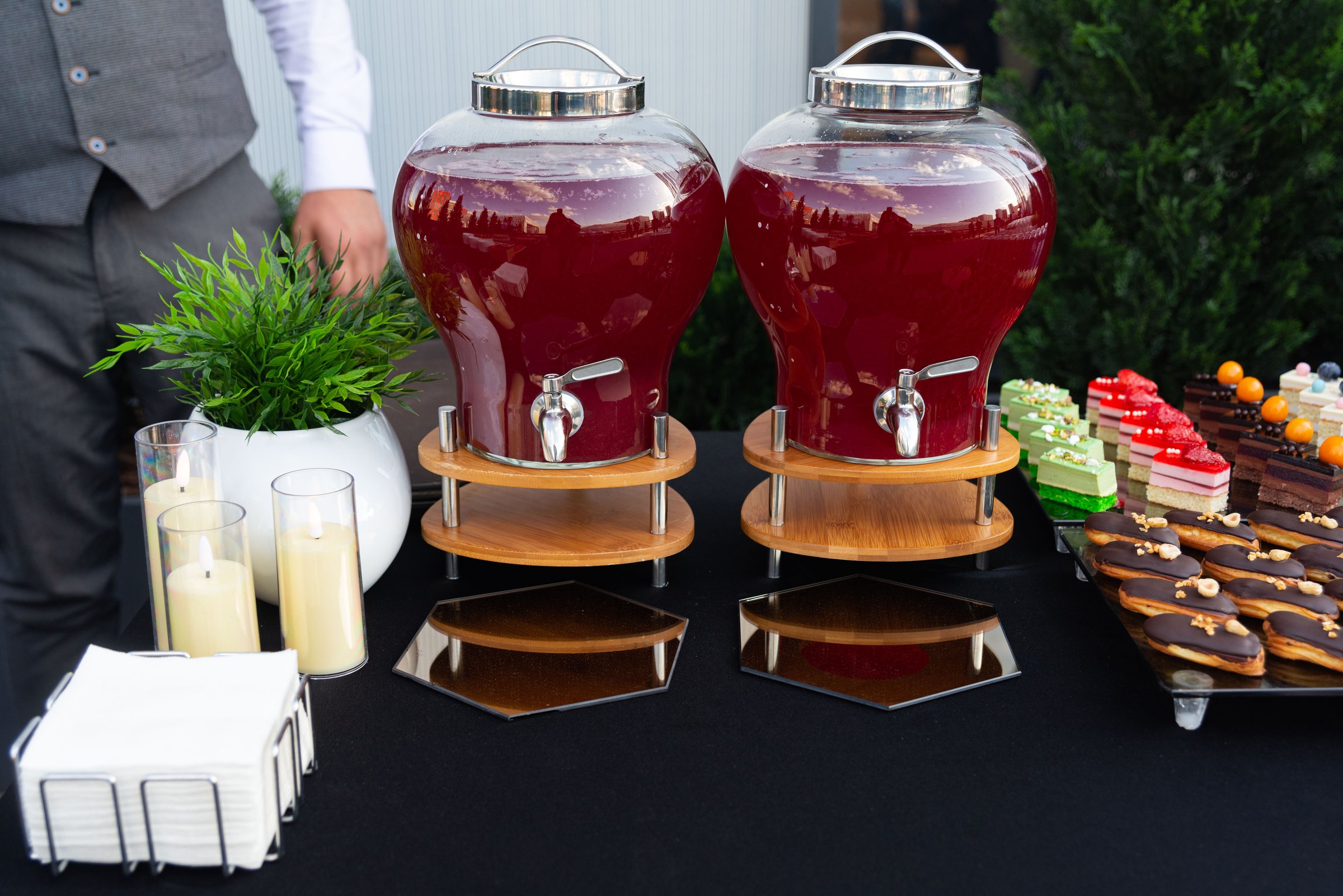 A stylish outdoor setup featuring two large drink dispensers filled with a vibrant red beverage. the table also includes an array of colorful desserts and candles, creating an inviting atmosphere.