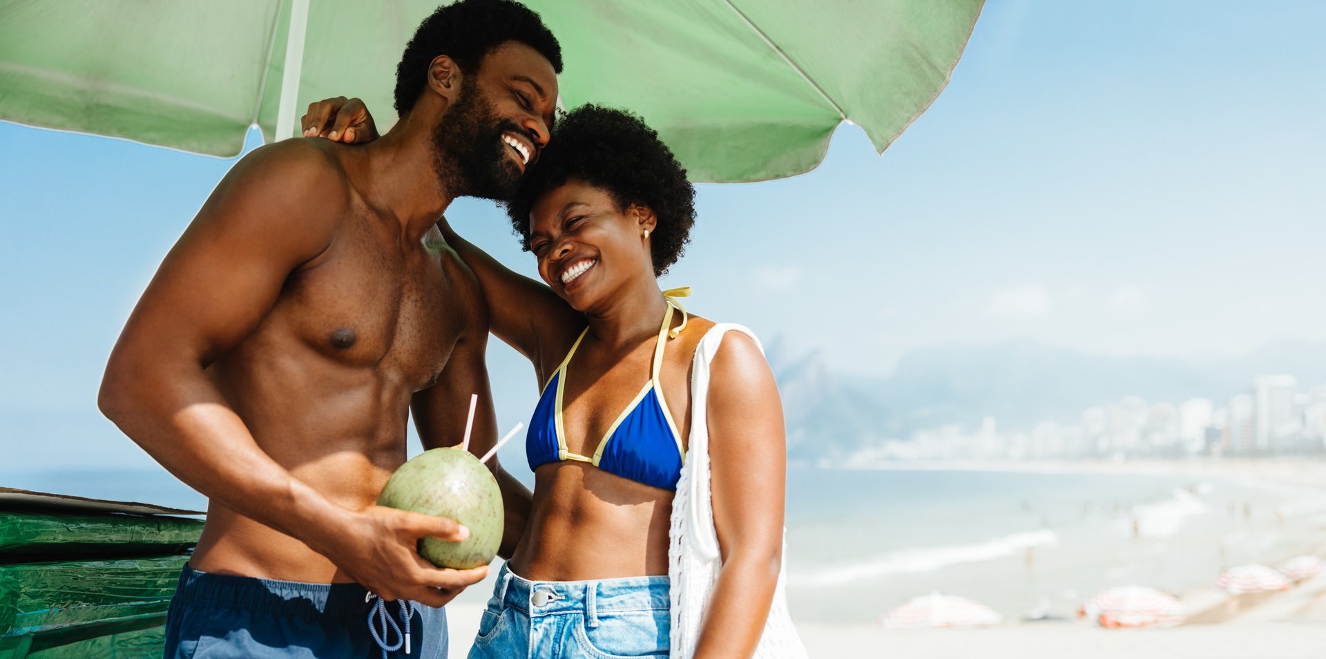 A happy African couple shares a joyful moment while relaxing on Ipanema Beach. The man is holding a fresh coconut as they both smile under a sun umbrella, with a picturesque backdrop of Ipanema Beach.
