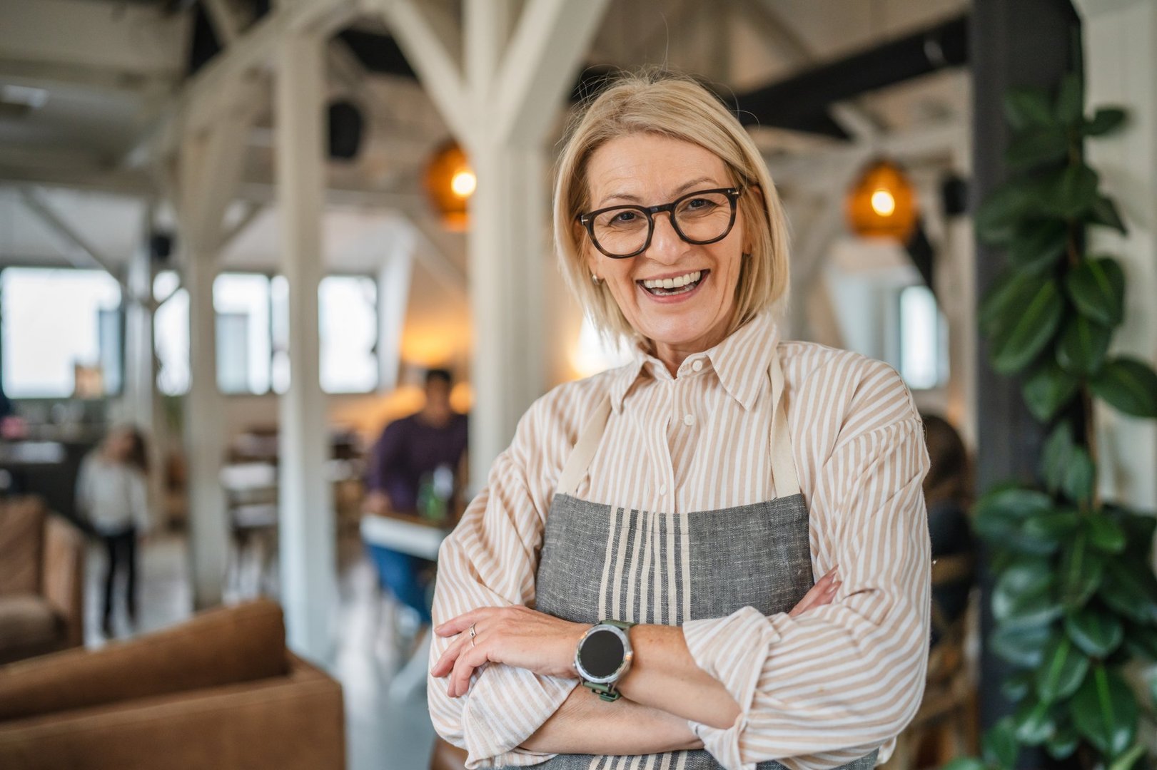 Portrait of a happy mature woman in apron stand at restaurant arm crossed, beautiful woman owner stand and smile at restaurant