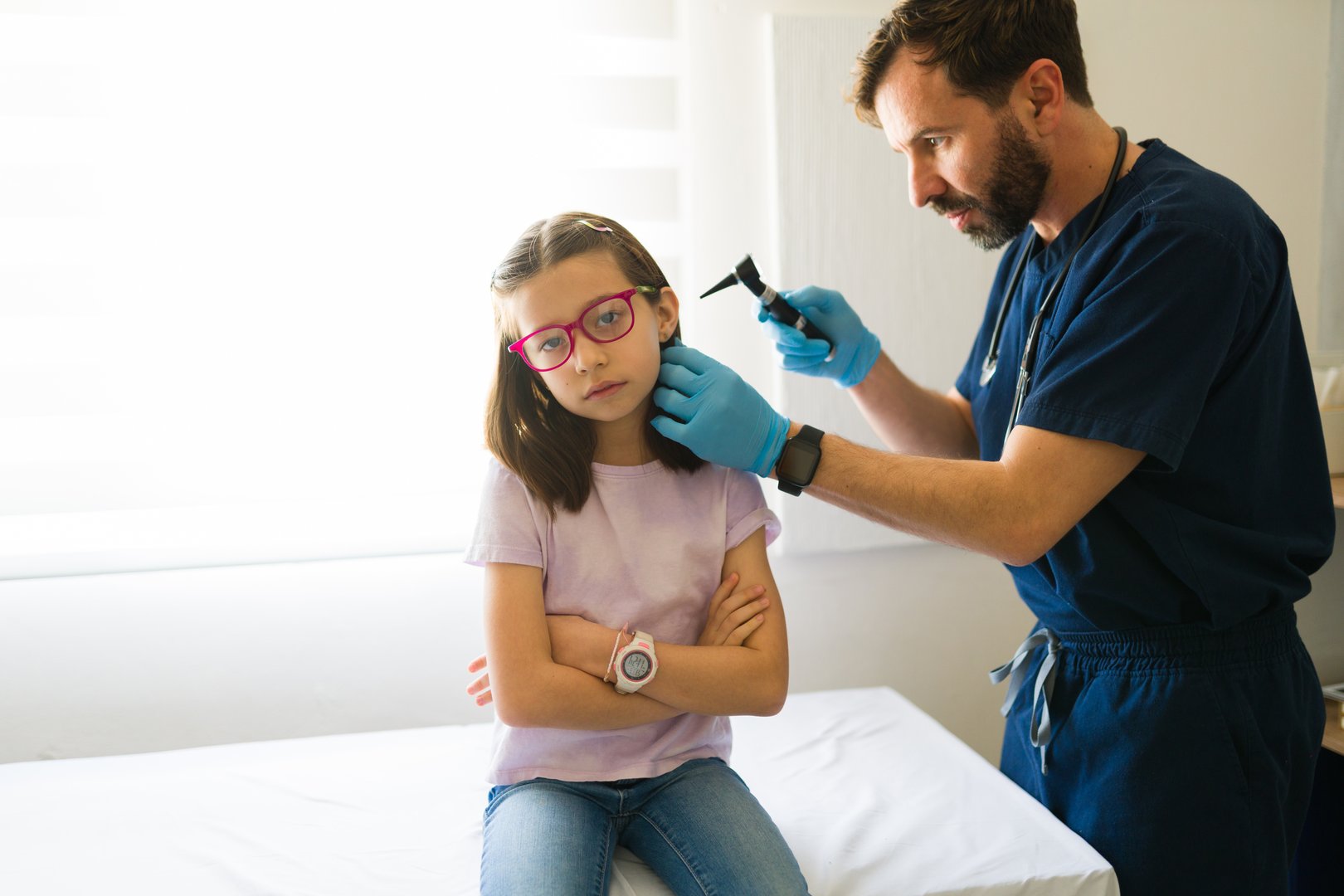 Pediatrician using an otoscope examining ear of a young girl sitting on examination table in doctor's office