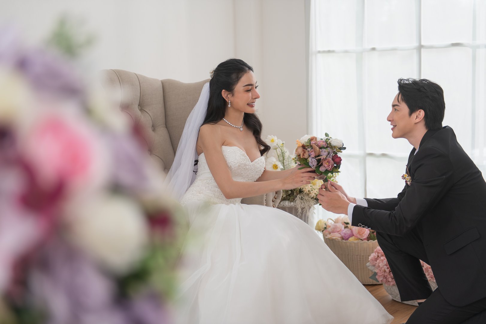 Groom kneels and offers a bouquet to his smiling bride in a white wedding dress, surrounded by floral decorations in a bright studio. Perfect romantic wedding moment.