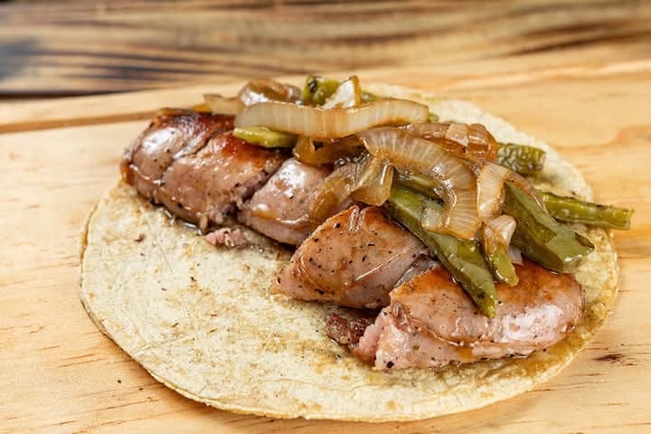 Taco with grilled meat, sautéed onions, and green peppers on a wooden board.