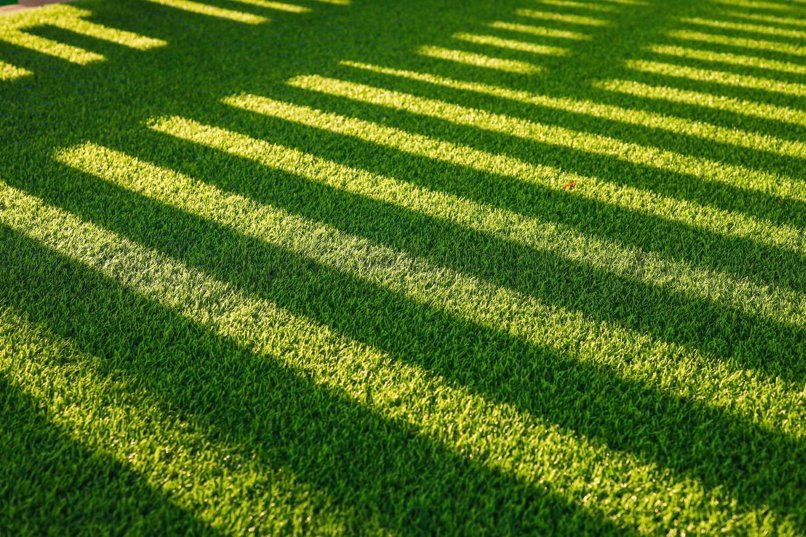 Close-up view of vibrant green artificial grass with dramatic shadows from the fence in sunlight. Minimalistic, abstract natural texture background.