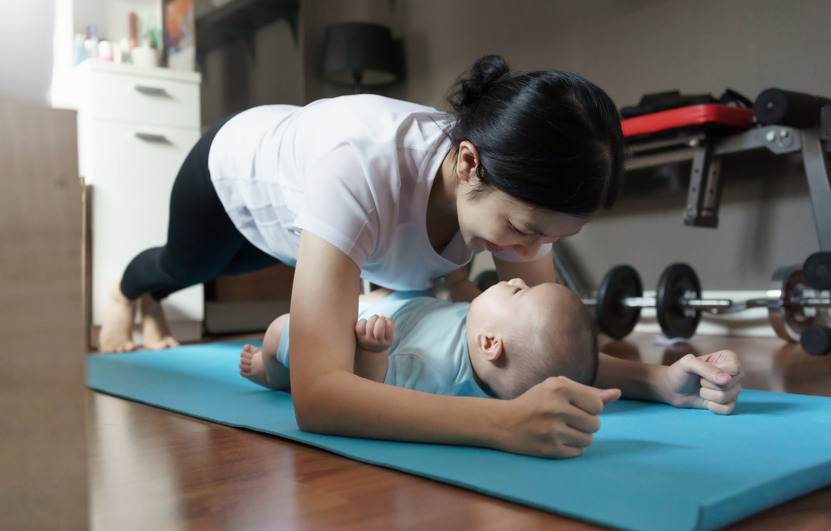 Happy Smiling Sporty Asian young mother doing plank exercise with her baby boy at home. Healthy Mom training and practicing yoga with her son on the floor in bedroom.
