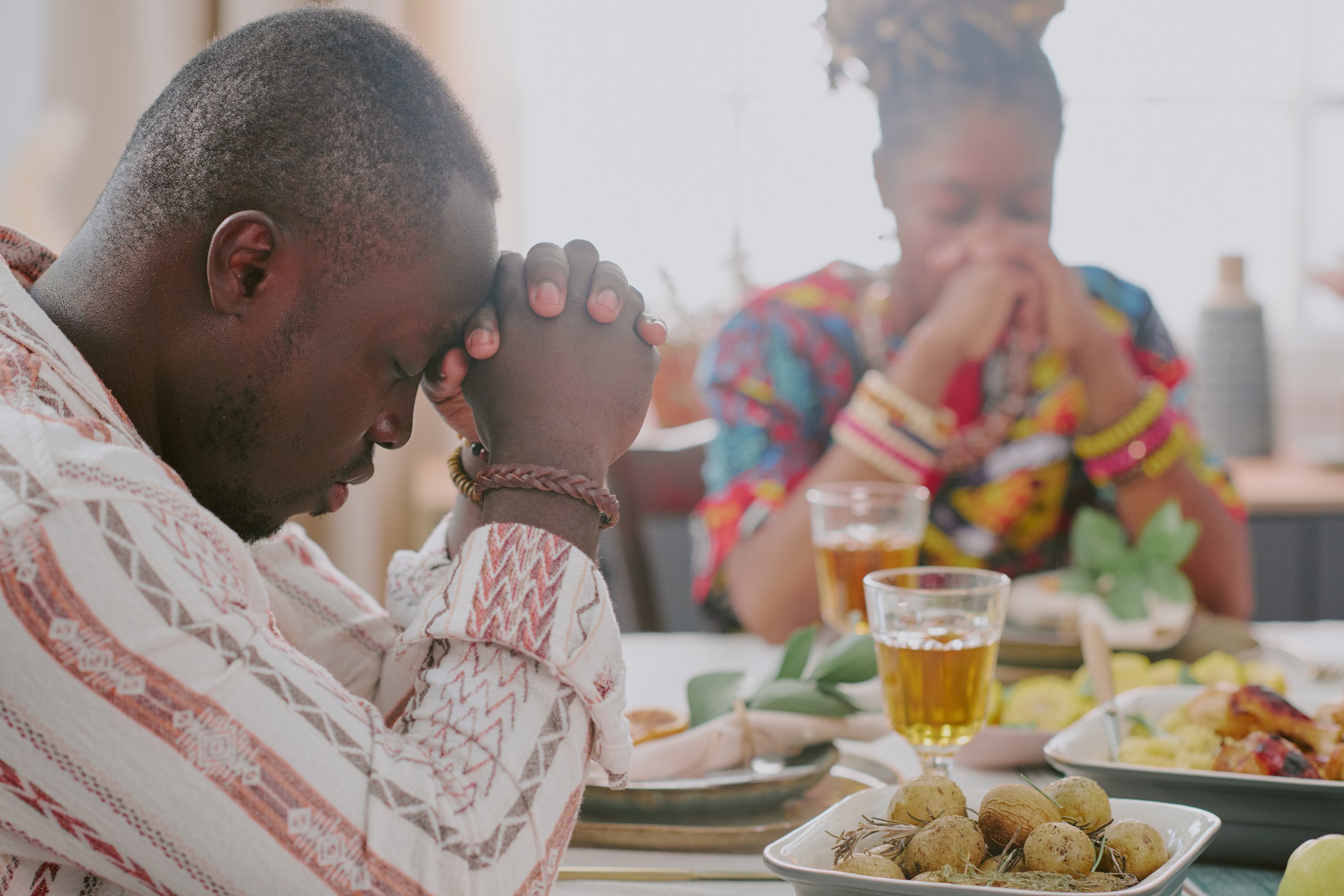 Black man and Black woman sitting at table praying before meal during Kwanzaa celebration, hands clasped and eyes closed, traditional dishes and decorations visible on table