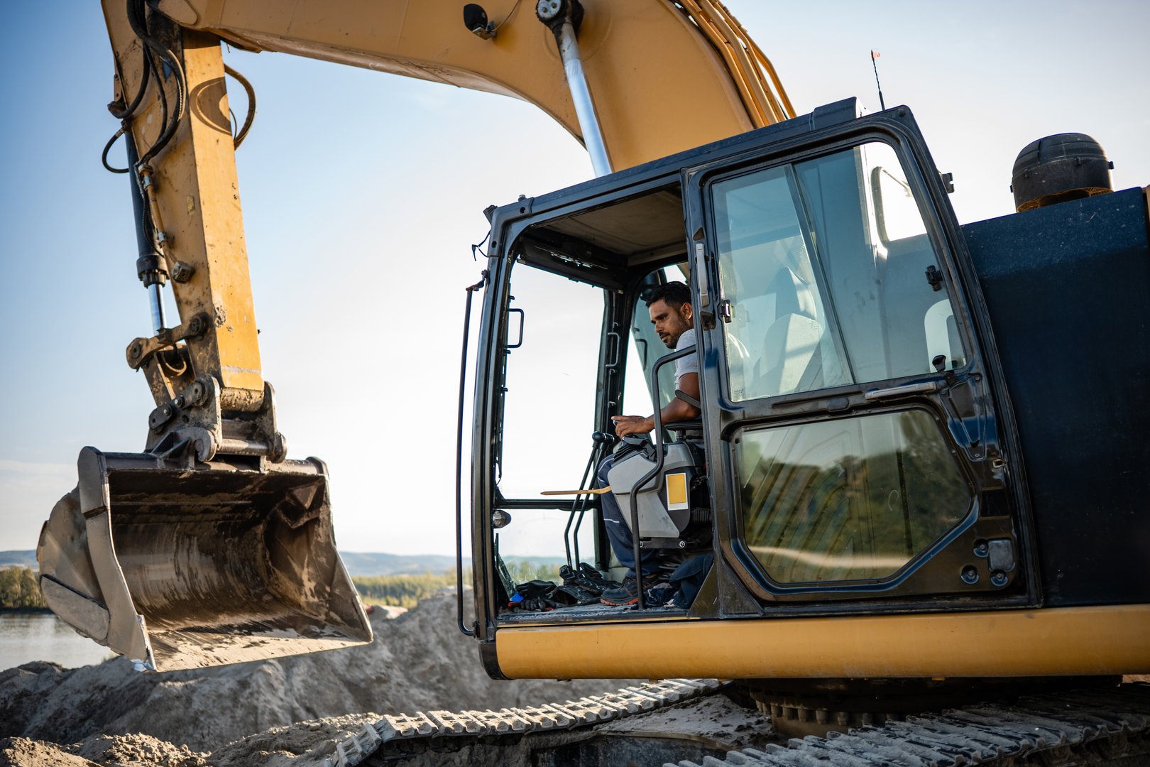 Construction worker operating a large excavator at a busy construction site beside a river, focused on earthmoving and infrastructure development