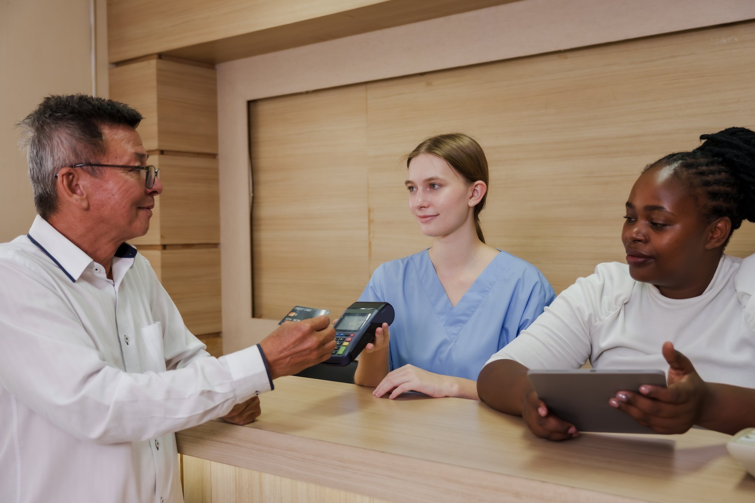 Senior asian male patient completing credit card payment at hospital reception desk with multiracial female medical staff assisting during billing process for healthcare services