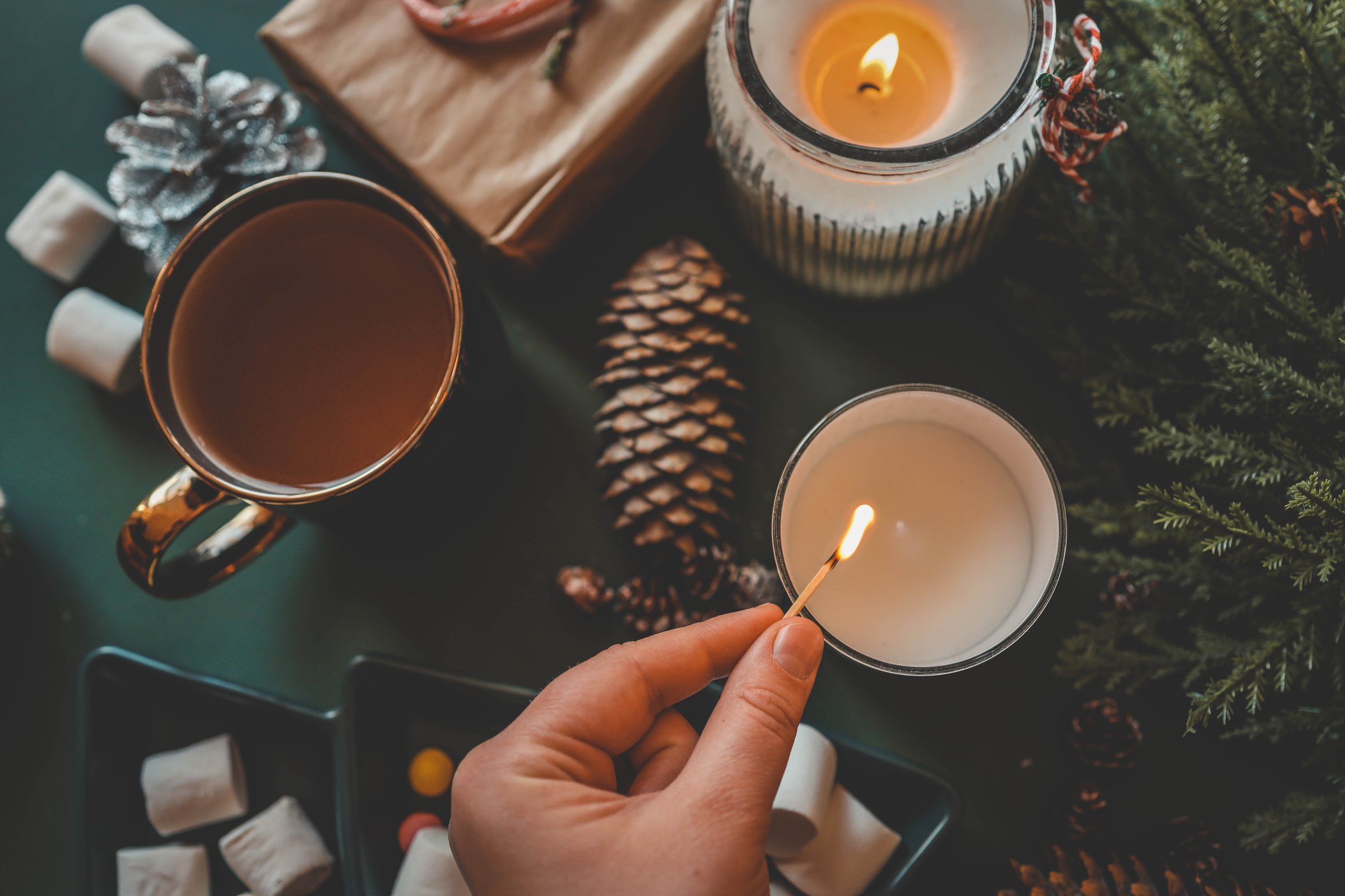 A hand lighting a festive candle with a matchstick, surrounded by pinecones, marshmallows, and evergreen branches, creating a warm and cozy Christmas atmosphere.