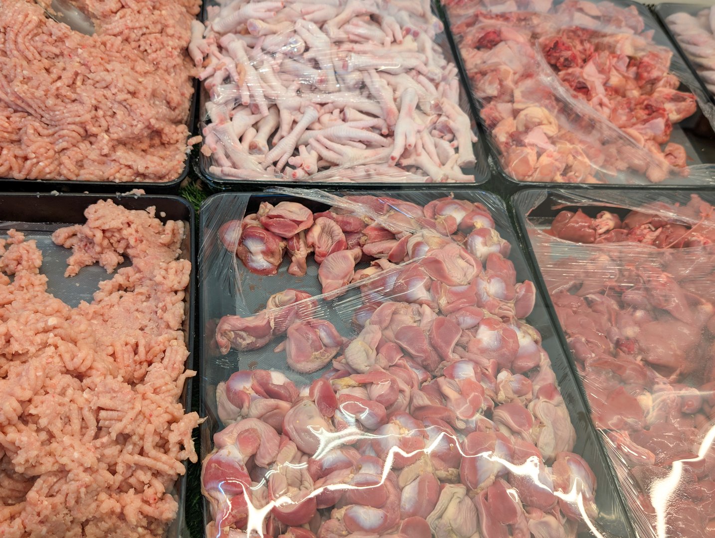 Various chicken meat cuts are displayed in a shop window. The selection includes ground chicken, chicken feet, gizzards, and livers arranged in trays
