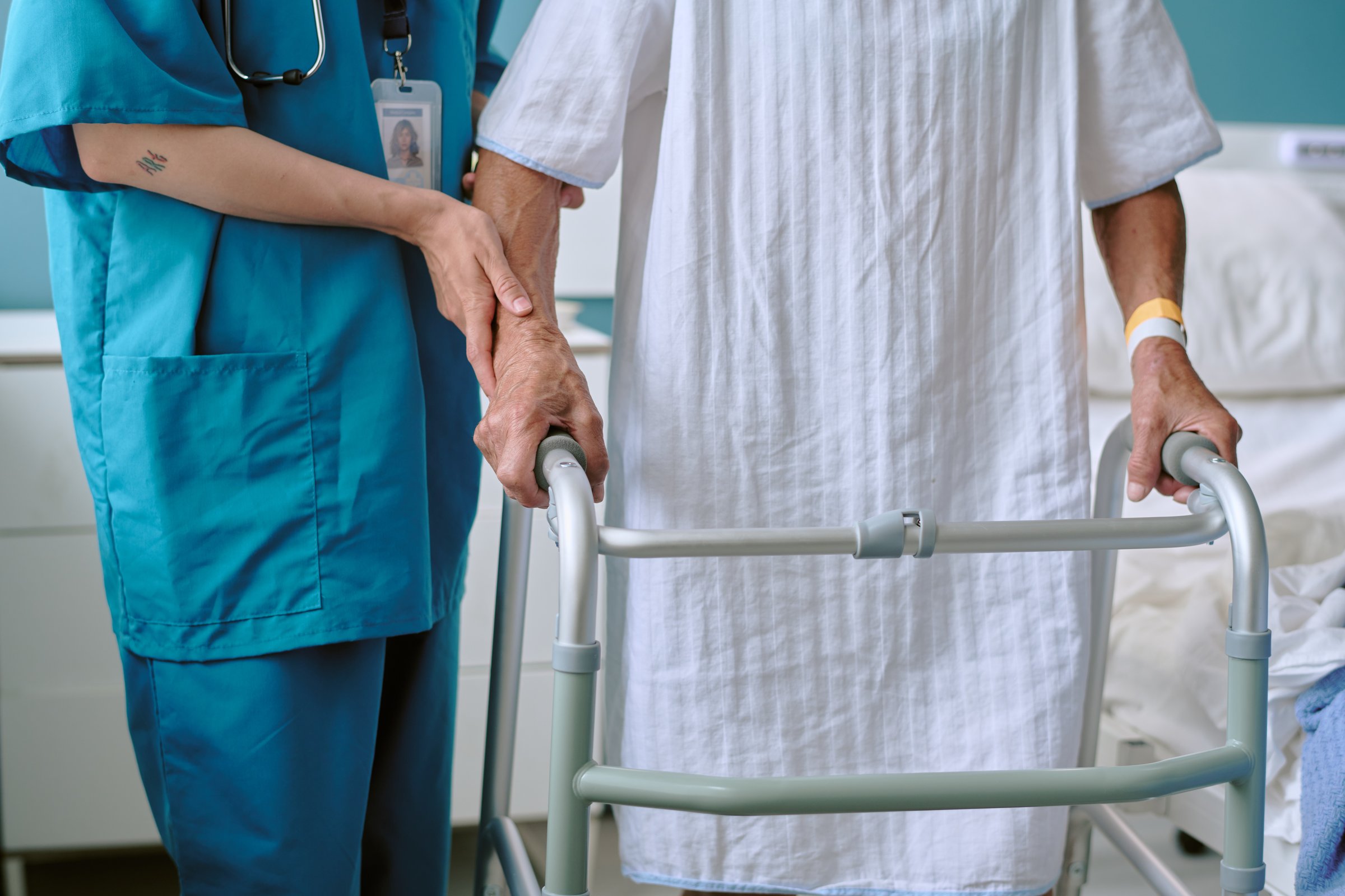 Senior Caucasian woman using walker for support while female healthcare worker assisting by holding arm, hospital bed visible in background, medical care scene focusing on mobility aid