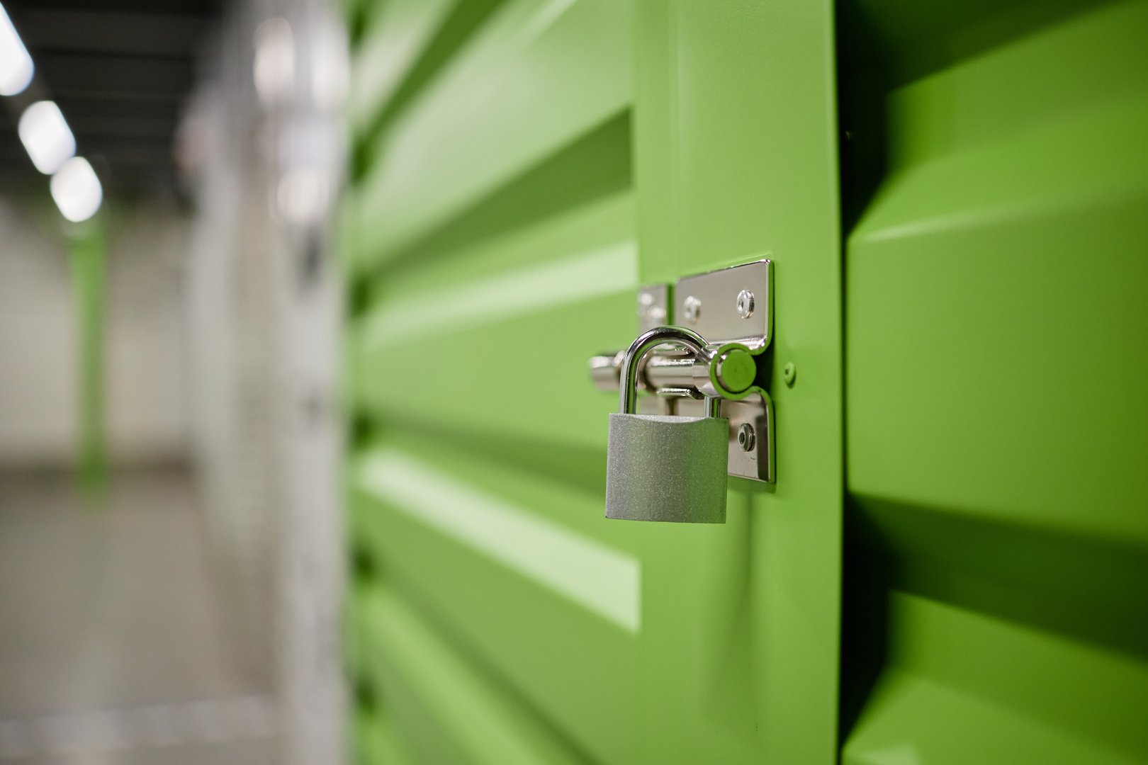 Metal padlock securing green storage unit door in indoor facility, focus on lock and latch mechanism with blurred corridor background, concept of security and self storage solutions