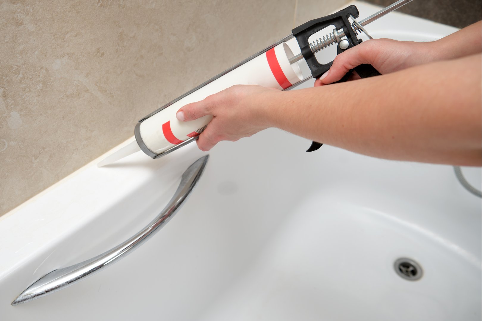 Hand applying sealant to bathtub corner using caulking gun. Home renovation and bathroom maintenance concept.