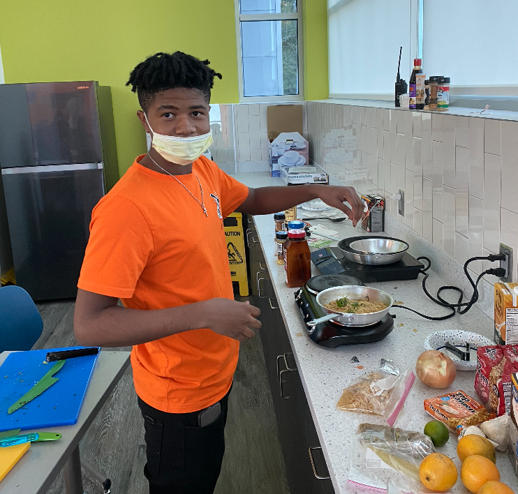 A person wearing an orange shirt cooks on a counter in a kitchen, with ingredients and kitchenware scattered around.