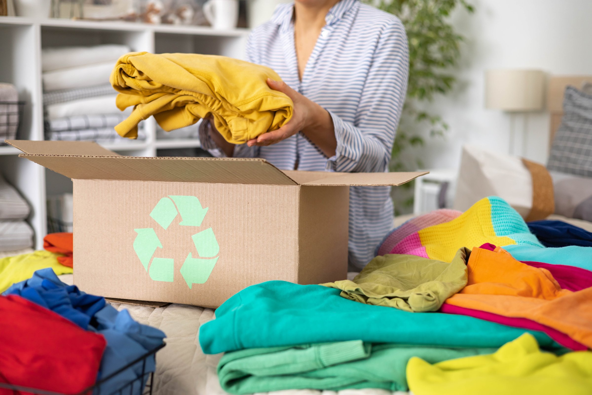 The concept of recycling clothes. A woman puts bright colored different clothes in a box with a recycle sign.
