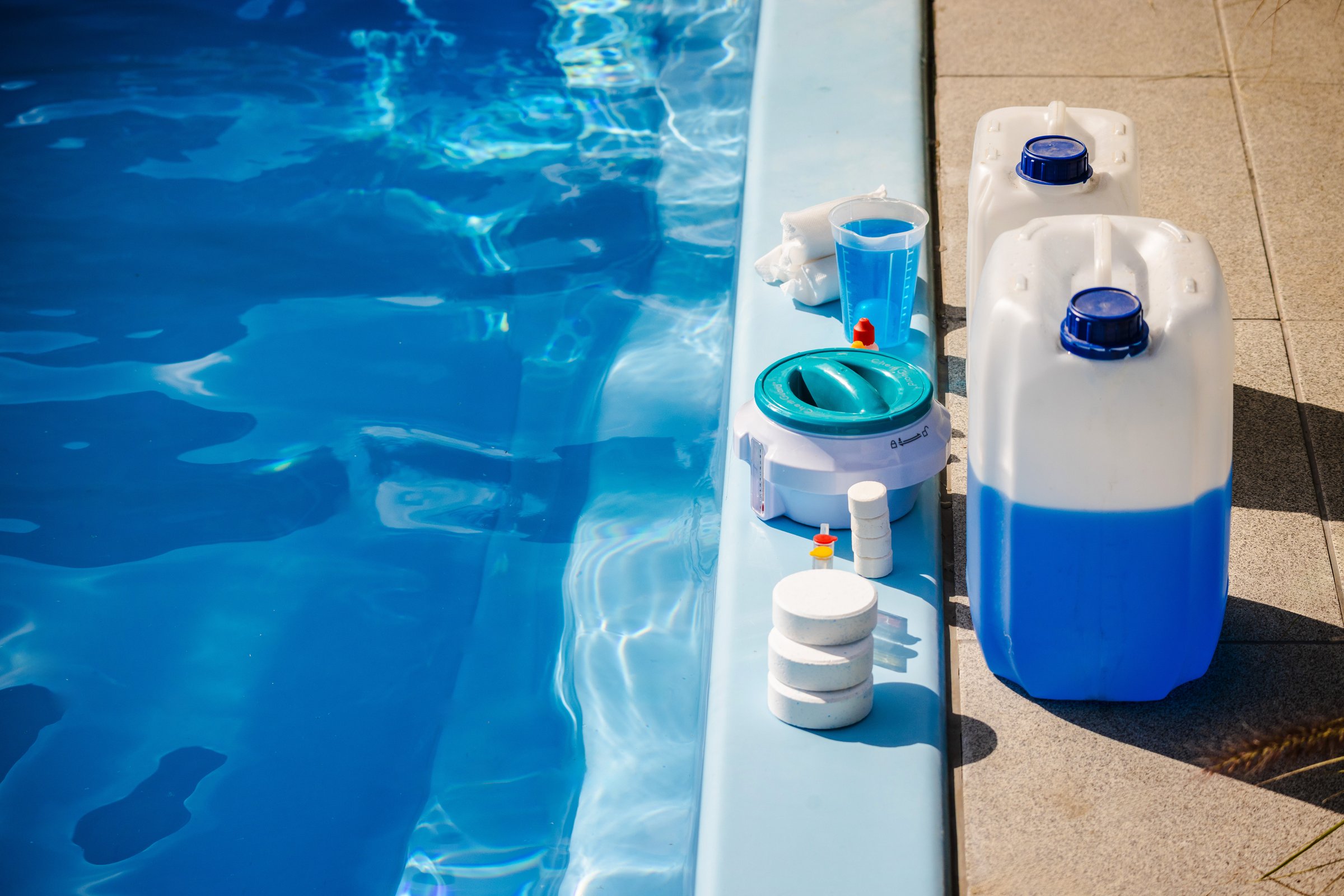 Swimming pool cleaning products including chemical canisters with blue liquid, chlorine tablets, floating dispenser, measuring cup and sanitizer bottles placed at pool edge. High quality photo