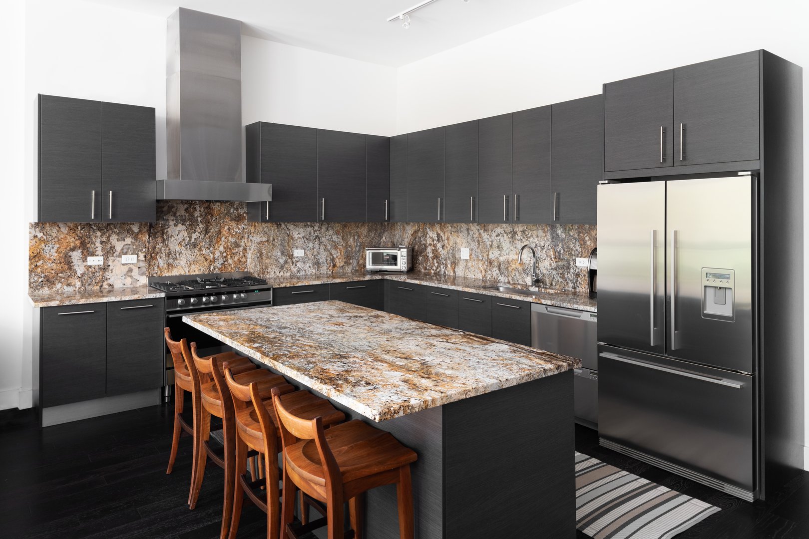 A modern kitchen with brown cabinets, granite countertops, bar stools sitting at the large island, and stainless steel appliances.