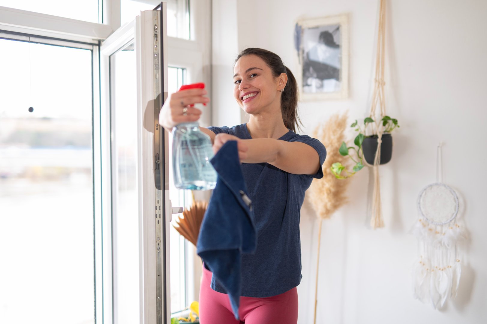 Woman spraying cleaner on window, smiling while doing housework