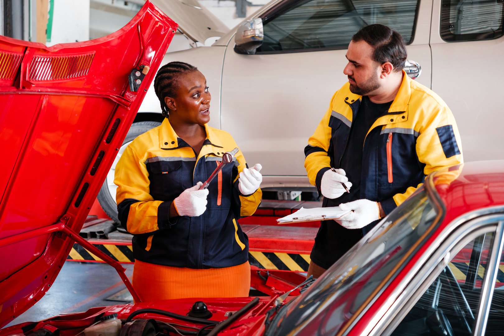 Diversity mechanic teamwork, an Indian man and an African woman in yellow and blue uniforms. A man inspects the car engine with his woman assistant. Automobile repairing service. Vehicle maintenance.