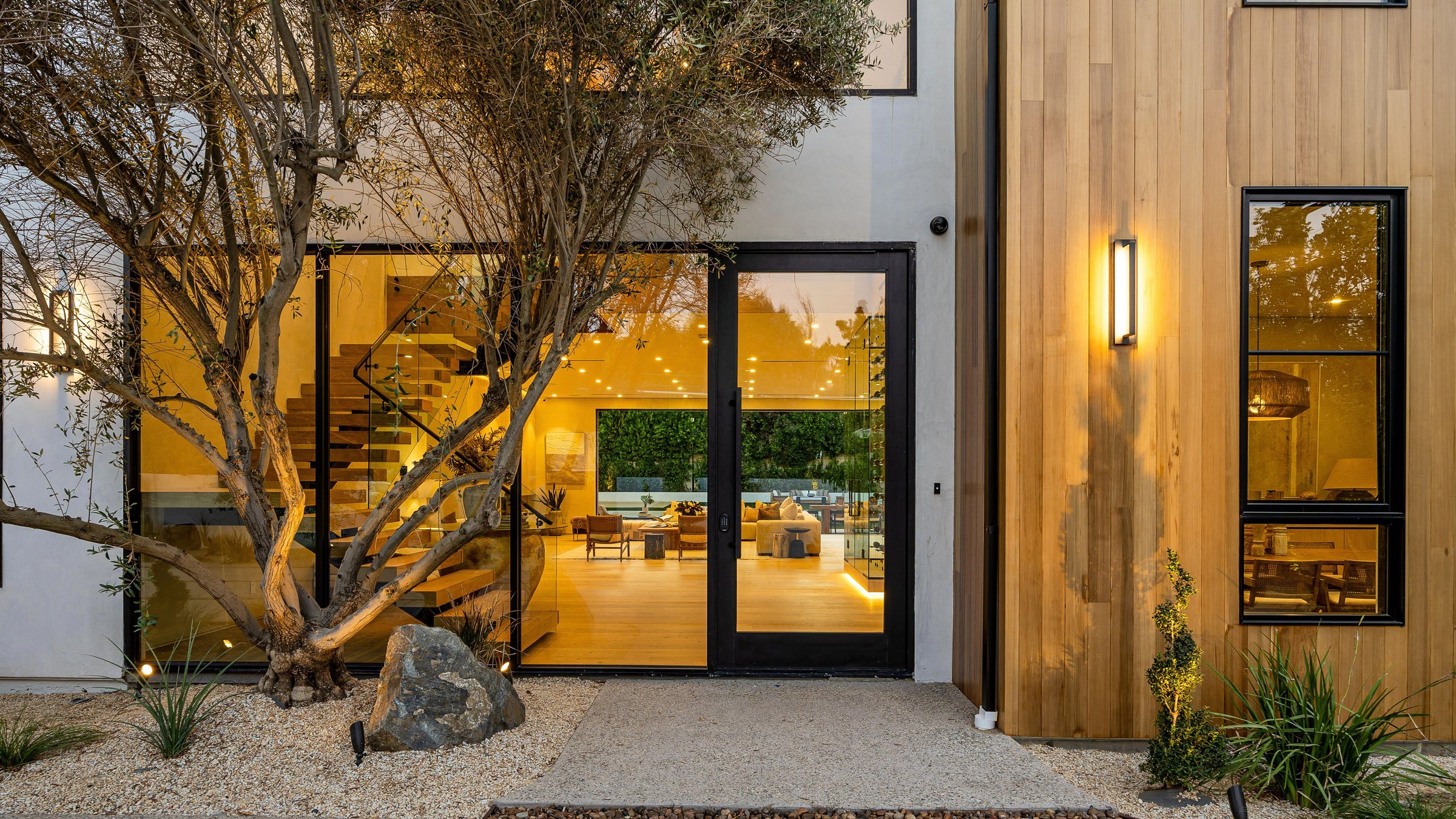 Modern home exterior with large glass doors, warm lighting, and a tree in the foreground, showcasing contemporary architecture.