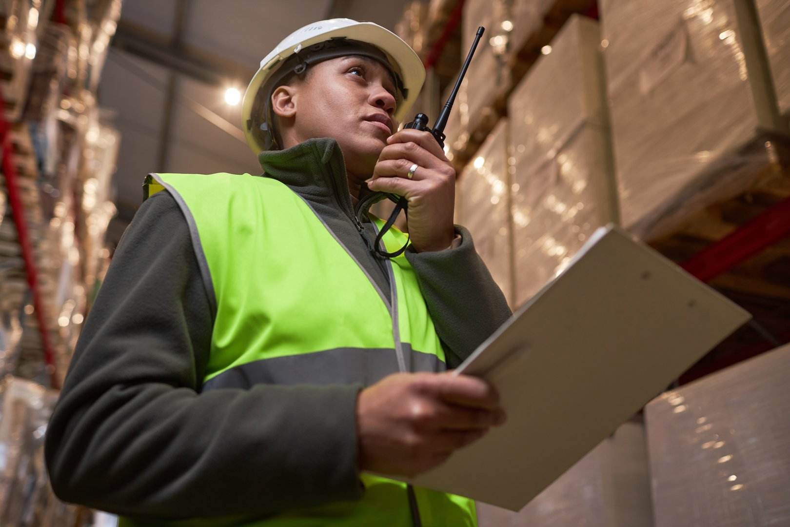 Low angle portrait of black woman wearing hardhat in storage warehouse and speaking to portable radio